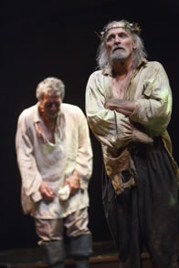 Lear in ragged white shirt and billowing brown pants, weeds stuck in his crown, his arms folded; behind, Gloucester in untucked white shirt and gray pants with knee boots stoops, crying